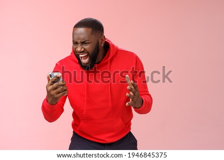 Angry furious african-american bearded guy lose control emotions shouting outraged annoyed smartphone display look phone insane stooping frowning rage, standing irritated pink background Royalty-Free Stock Photo #1946845375