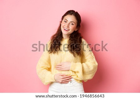 Young smiling woman touching her stomach with relieved, happy face, feeling good after eating yoghurt or medicine from painful cramps, standing against pink background Royalty-Free Stock Photo #1946856643