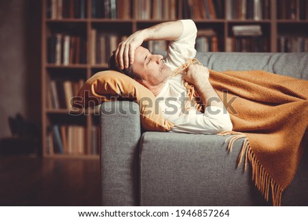 An adult man lies on the couch at home against the background of a bookcase with symptoms of the disease. Royalty-Free Stock Photo #1946857264