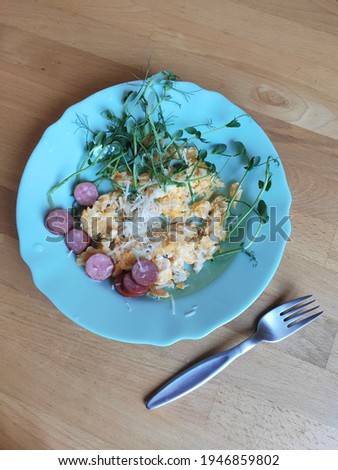 Top view photo of a blue plate with scrambled eggs, sausage, grated cheese and micro greens - pea sprouts, - and a fork lying next to it on the table. Healthy breakfast concept.