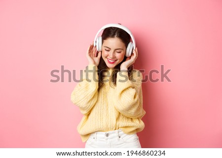 Portrait of attractive brunette girl enjoying listening music, dancing and smiling pleased, standing against pink background
