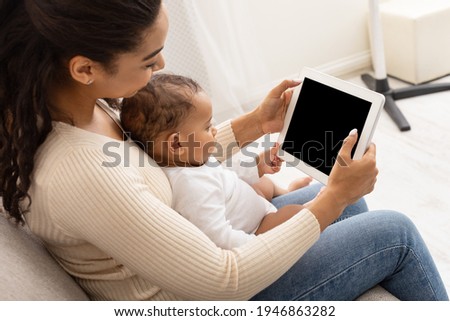 African American Mom With Toddler Baby Using Digital Tablet With Blank Screen Sitting On Sofa At Home. Side View Of Mom And Infant Using Computer Together. Application Advertisement. Mockup