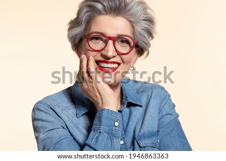 Close up portrait of attractive senior woman in modern red eyeglasses. Happy older female teacher with trendy haircut and toothy smile on beige background. Eyesight problem and stylish eyewear Royalty-Free Stock Photo #1946863363