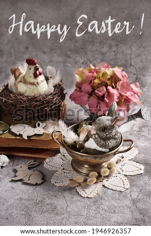 Easter card with a hen figurine in the nest and cute bunny in metal bowl on grunge gray background with greeting text