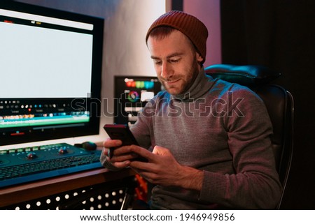 Attractive Male Video Editor Works with Footage or Video on His Personal Computer and having a break communicating on his smartphone. He Works in Creative Office Studio or home. Neon lights