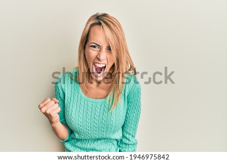 Beautiful blonde woman wearing casual winter sweater angry and mad raising fist frustrated and furious while shouting with anger. rage and aggressive concept.  Royalty-Free Stock Photo #1946975842