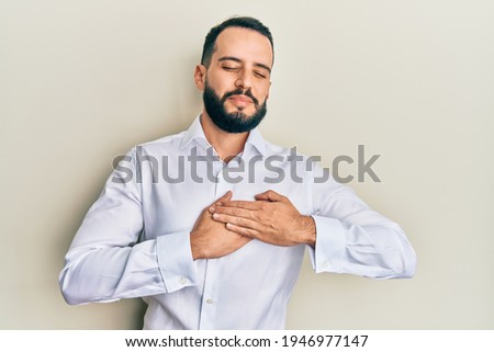Young man with beard wearing business white shirt smiling with hands on chest, eyes closed with grateful gesture on face. health concept. 