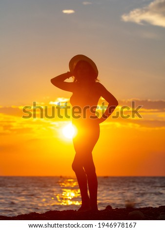 Silhouette of a girl in a swimsuit with a hat near the sea at sunset.