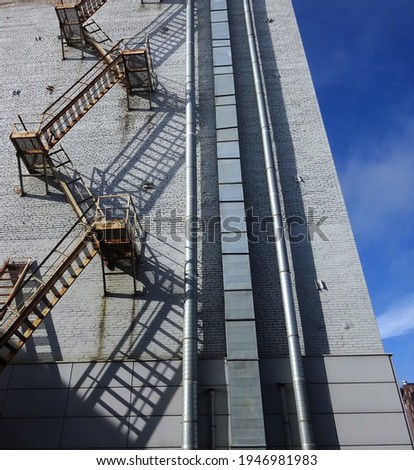 An upwardly directed brick wall with ventilation pipes, an iron fire escape and a variety of deep shadows. Industrial zone concept. Production building background with copy space. Vertical photo.

   