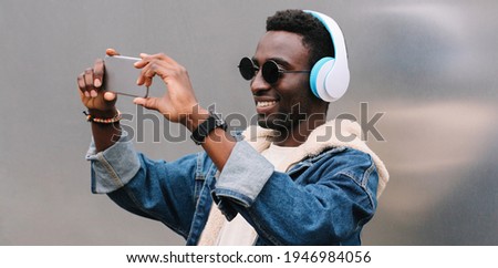 Portrait of happy smiling young african man taking selfie by smartphone listening to music in headphones on a city street