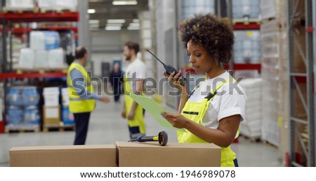 African female warehouse manager holding clipboard and using walkie-talkie while checking inventory. Black woman supervisor with clipboard and portable radio in storehouse Royalty-Free Stock Photo #1946989078