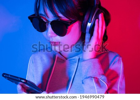 ํYoung woman​ in headphones listening​​ to music​ or playing game with smartphone on neon light background.
