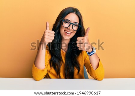 Beautiful brunette young woman wearing glasses and casual clothes sitting on the table success sign doing positive gesture with hand, thumbs up smiling and happy. cheerful expression.