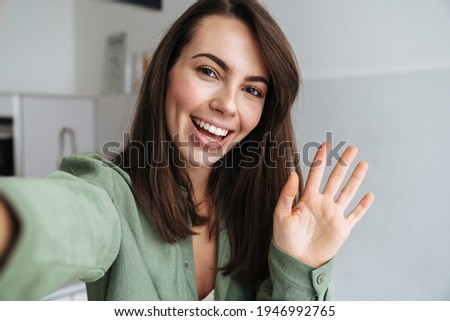 Young happy woman waving hand while taking selfie photo at home kitchen