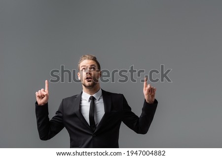 astonished businessman looking up and pointing with fingers isolated on grey