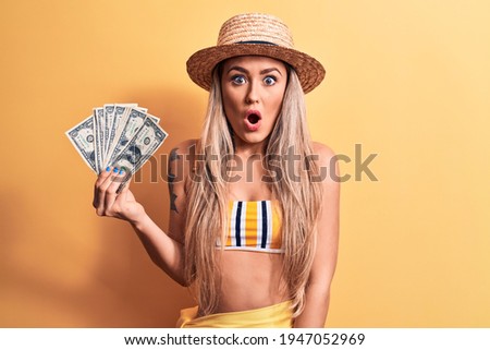 Young beautiful blonde woman wearing bikini and hat holding bunch of dollars banknotes scared and amazed with open mouth for surprise, disbelief face