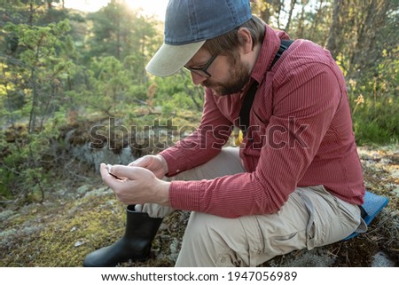 Man is resting after a long walk, he looks at his smartphone, sitting on a stone covered with moss, in the forest.