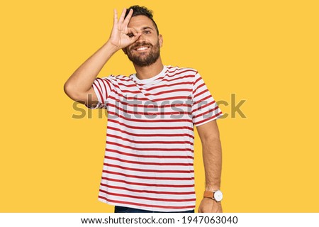 Handsome man with beard wearing striped tshirt smiling happy doing ok sign with hand on eye looking through fingers 