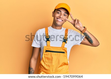 Young handsome african american man wearing handyman uniform over yellow background doing peace symbol with fingers over face, smiling cheerful showing victory 