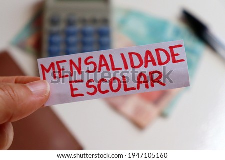 Translation: Tuition (mensalidade escolar). | Hand holding note with mensalidade escolar (tuition) written and calculator, Brazilian money and wallet in the background. School tuition fees in Brazil.