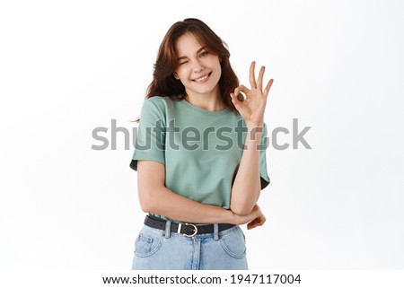 Beautiful teenage girl student, smiling and showing okay sign, likes item, approve and praise good choice, guarantee quality, recommend product, standing against white background