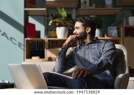 Young thoughtful smart indian professional business man executive looking away relaxing sitting on chair in modern office lobby with laptop, thinking of new ideas, dreaming of success, planning. Royalty-Free Stock Photo #1947138829
