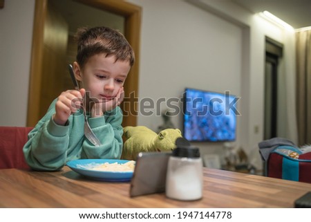 Cute little boy eating dinner and looking at the smartphone in the kitchen. Young small kid staring and watching cartoon on smartphone while eating food on dining table
