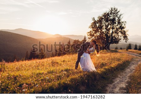 Beautiful newlyweds couple hugging in mountains at sunset. Bride and groom walking in summer Carpathians among flowers. Wedding