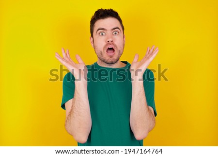 Surprised terrified  young handsome caucasian man wearing green t-shirt against yellow background Gestures with uncertainty, stares at camera, puzzled as doesn't know answer on tricky question, People