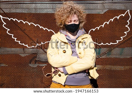 Fashionable curly blonde woman girl with closed eyes 30 plus years in a black protective mask with chalk painted wings on an rusty background. COVID-19 coronavirus. Health people. Woman modern fashion