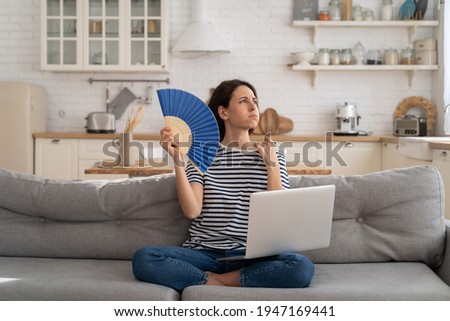 Tired millennial woman suffering from heatstroke flat without air-conditioner, waving blue fan, sitting on couch at home, working on laptop computer. Overheating, high temperature, hot summer weather. Royalty-Free Stock Photo #1947169441