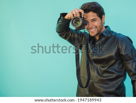 Young Latino man holds a camera on an Aztec green background with copy space. Focus on the face