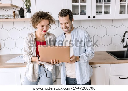 Happy family couple are standing at the kitchen at home, doing online shopping or chatting with friends. Joyful stylish husband and wife using laptop, watching funny videos or photos, smiling