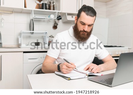 Photo of young man with beard sitting at home and making some notes in planner.