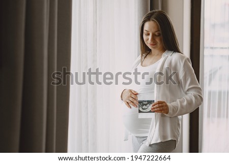 Pregnant woman stands by the window and looks at photo