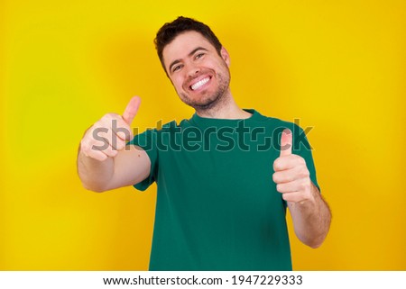  young handsome caucasian man wearing green t-shirt against yellow background approving doing positive gesture with hand, thumbs up smiling and happy for success. Winner gesture.