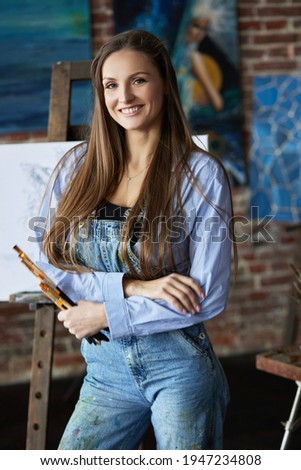 Portrait of young smiling female artist with her artworks in art studio. Painter holding art brushes. Creative process, relaxation, leisure, hobby, stress management.