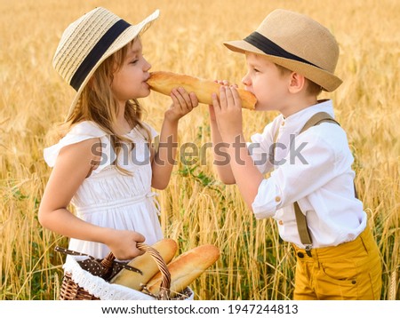Little boy and girl in hats stand in a wheat field with a basket of bread and share one loaf between two, bite a loaf together. The child eats in the fresh air. Walk and picnic of friends on nature. 