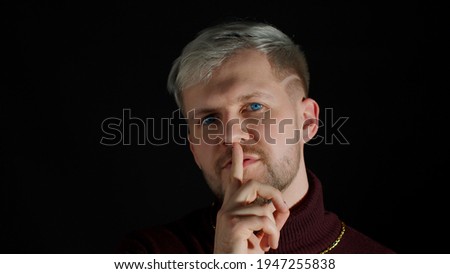 Strict serious man asking to stay calm, keep silence or secrecy with finger on lips gesture, mysterious confidential information on black background. Facial expression of trendy guy. Shh, be quiet
