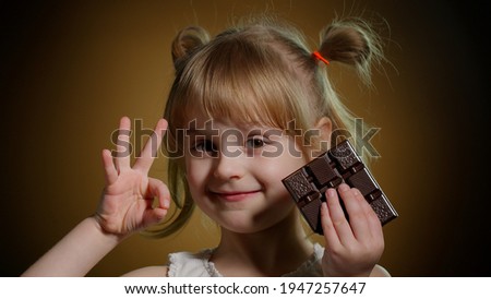 Close-up portrait of teen child kid with milk chocolate bar showing thumb up gesture, ok sign. Addiction of sweets, candies. Satisfied young blond girl with ponytails smiling on dark brown background
