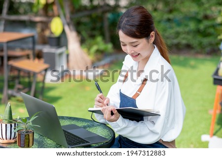 Beautiful Asian woman record the data into notebook in front of laptop and stay in home garden with happy emotion.
