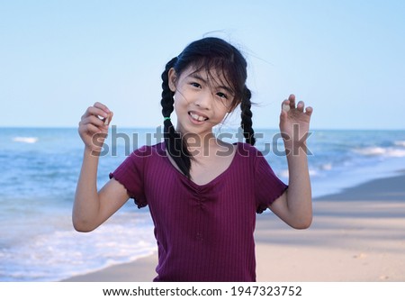 A cute young Asian girl is showing off her seashells that she had collected along a white sandy beach by the sea, with beautiful calm clear water in the background. 