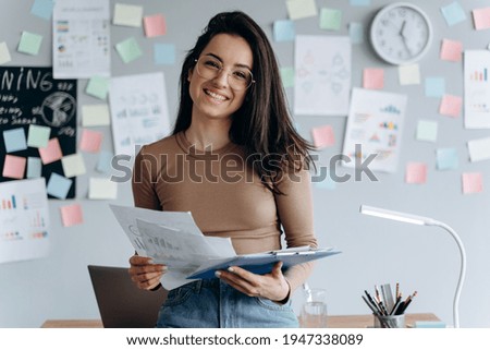 Beautiful, young business lady holds important documents in her hands, she reviews them. Smiling, charming girl with glasses works in a modern office.