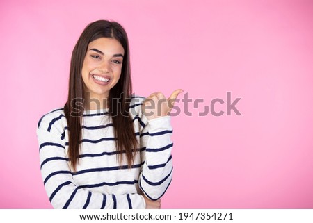 Horizontal shot of prettty young joyful female points with fore finger away, indicates copy space on blank wall, isolated over pink background. Check this out. 