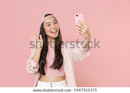 Smiling young asian woman girl in casual clothes cap posing isolated on pastel pink wall background. People lifestyle concept. Mock up copy space. Doing selfie shot on mobile phone greeting with hand