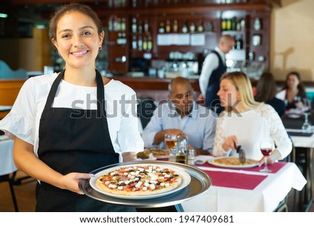 Hospitable latin american waitress holding plate with appetizing pizza, meeting guests in cozy pizzeria Royalty-Free Stock Photo #1947409681
