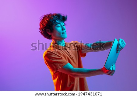 Studing, working. Young joyful man using tablet isolated on multicolored background in neon light. Concept of human emotions, facial expression, youth culture, digital life. Copy space for ad.