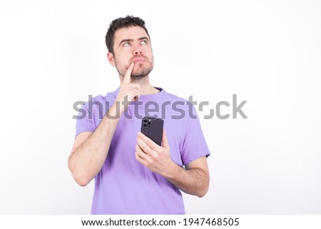 young handsome caucasian man wearing purple t-shirt against white background thinks deeply about something, uses modern mobile phone, tries to made up good message, keeps index finger near lips.