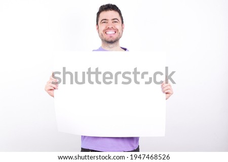 Happy young handsome caucasian man wearing purple t-shirt against white background Holding Empty Paper Board Advertising Offer Text Standing. Autumn Advertisement Banner.