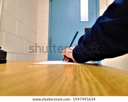 A man writing on paper with a blue pen and his arm stretched out. A blue capped pen is in his hand and his paper is a blank canvas to write on in this empty office space.